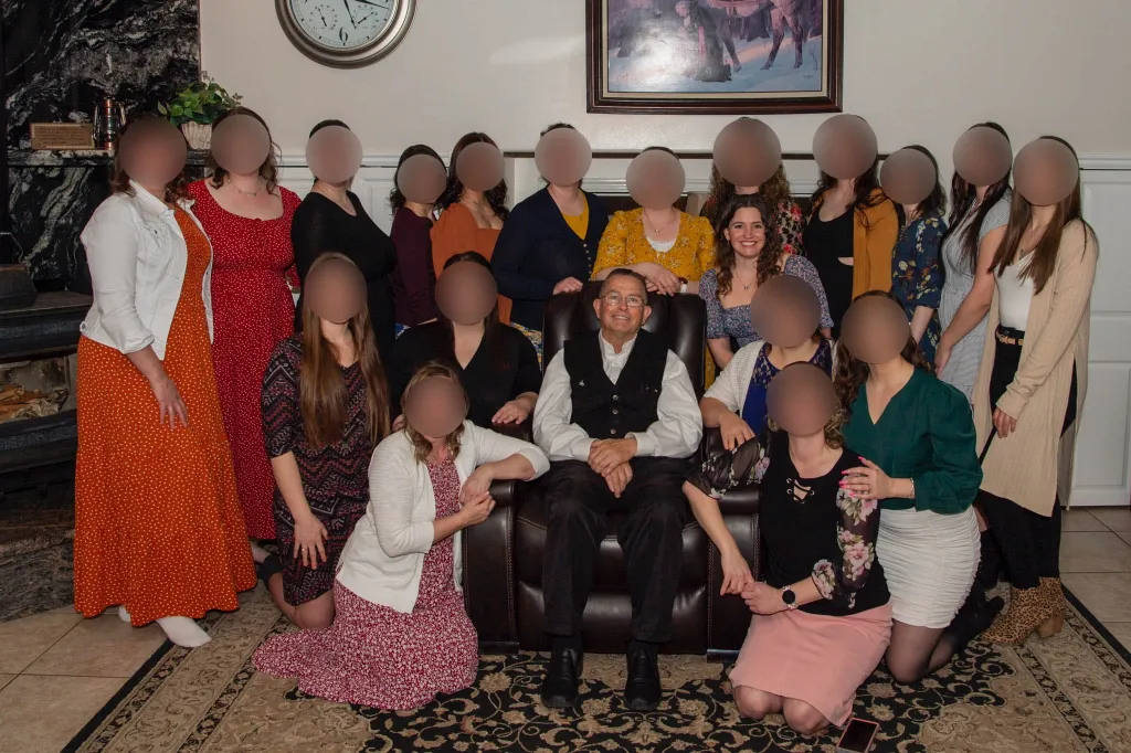 A man sitting in a chair surrounded by 19 women, with faces blurred out, including Janet, one of his 44 children.