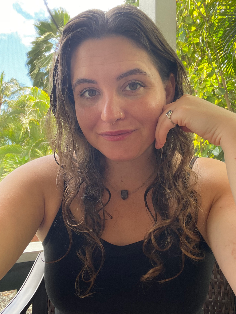 A woman with long wavy brown hair, wearing a black tank top, with her hand resting on her face.