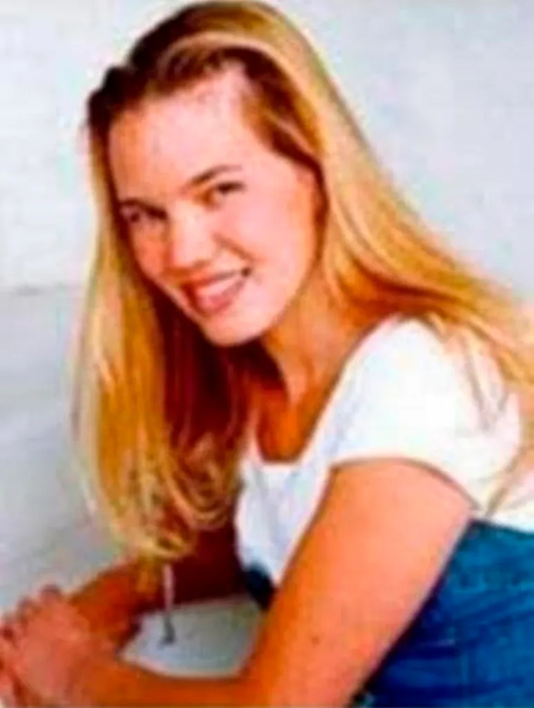 Kristin Smart, a white woman with long blonde hair, smiles faintly while wearing a white shirt and blue jeans.
