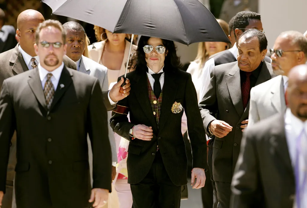 Michael Jackson walks out of a courthouse with his father Joseph Jackson, surrounded by other men and holding an umbrella.