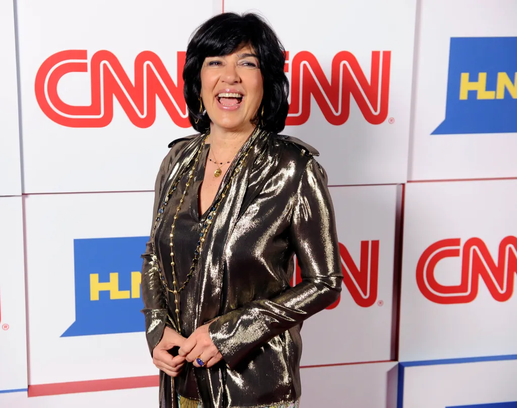 Christiane Amanpour in a gold lamé top smiling in front of a white CNN and HLN branded backdrop.