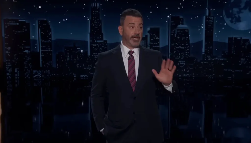Jimmy Kimmel gestures with his hand as he speaks to the camera in front of a backdrop of a city skyline at night.
