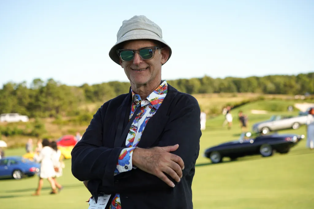Bob Rubin in a bucket hat, sunglasses, and a colorful shirt, standing on a grassy field with classic cars in the background.