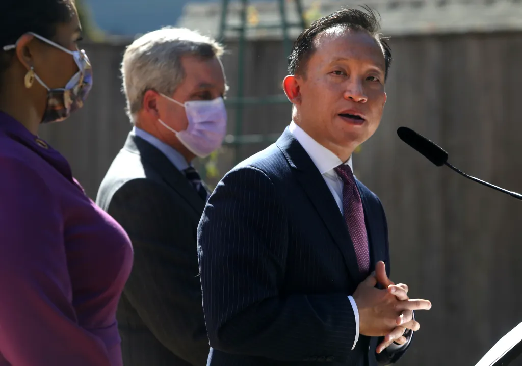 San Francisco City Attorney David Chiu speaking at a news conference, with Mayor London Breed and former City Attorney Dennis Herrera in the background.