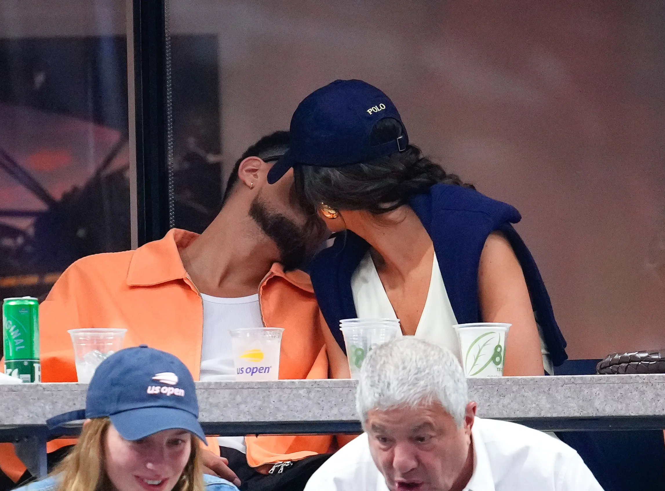 Devin Booker with a beard and an orange jacket, kisses Kendall Jenner, who wears a navy Polo hat and blue jacket, at the US Open.