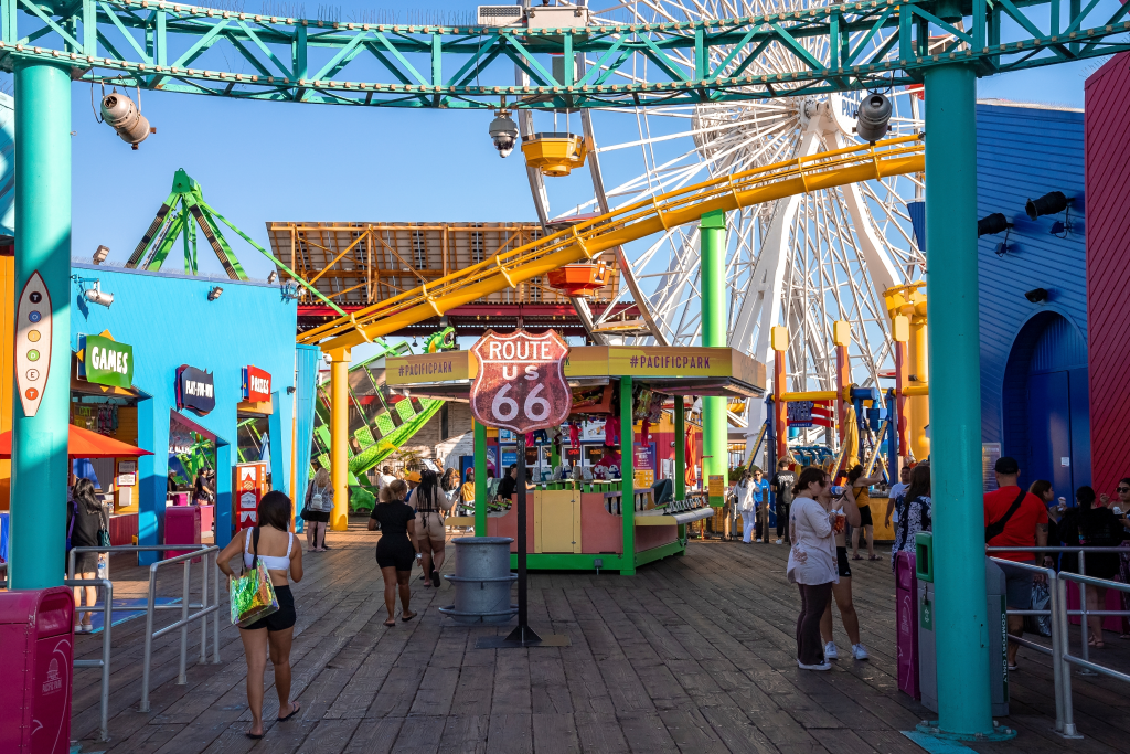 Tourists walking through Pacific Park on Santa Monica Pier, past a Route US 66 sign and various shops and rides.
