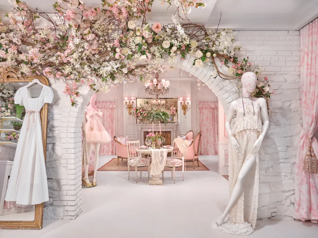 A white brick archway decorated with pink and white flowers leads to a room with a dining table and pink chairs, with a white dress hanging on a mirror to the left and mannequins in dresses on both sides of the archway.