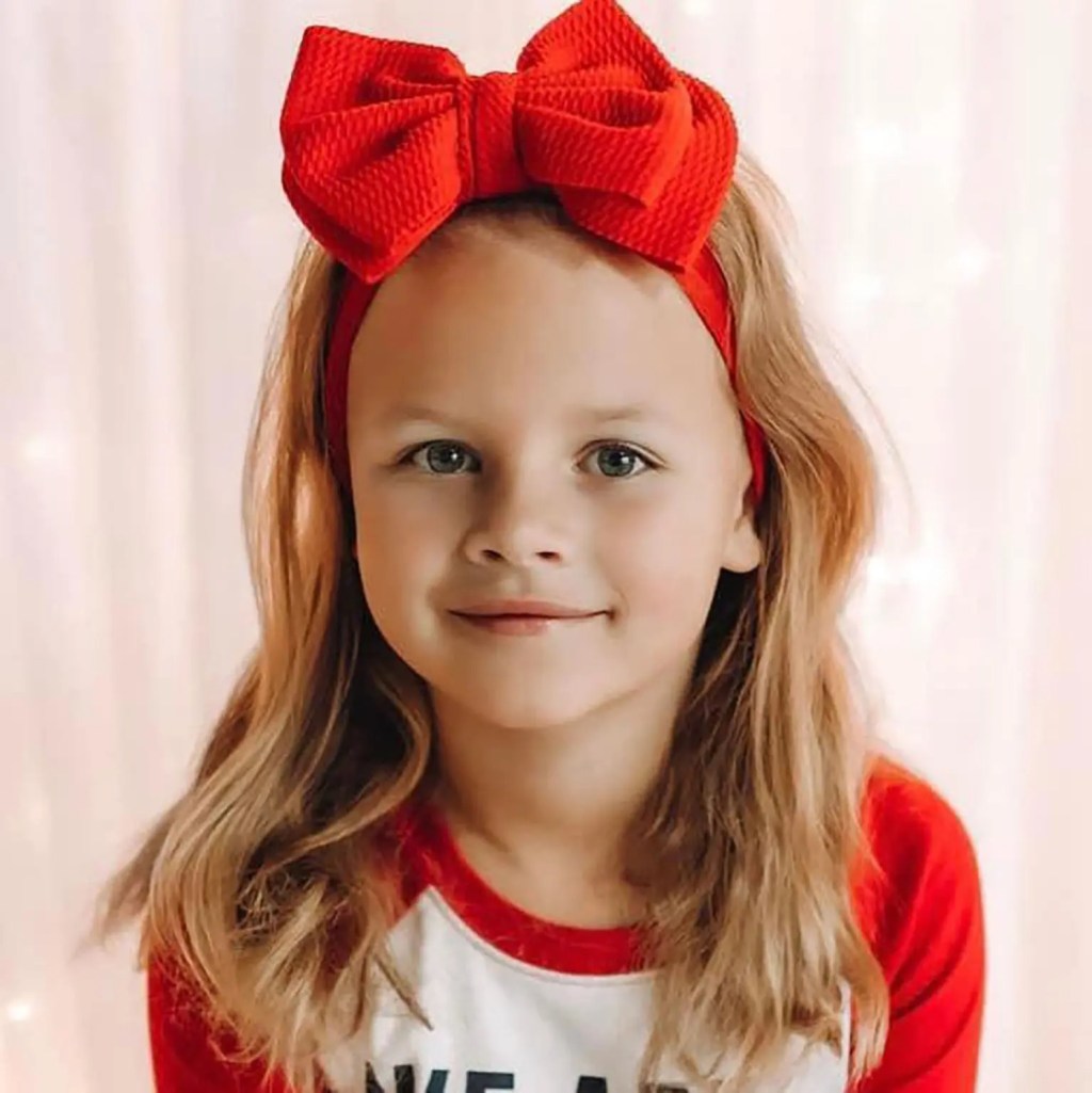 A young girl, Athena Strand, with long blonde hair and a red headband with a large red bow, smiles faintly.