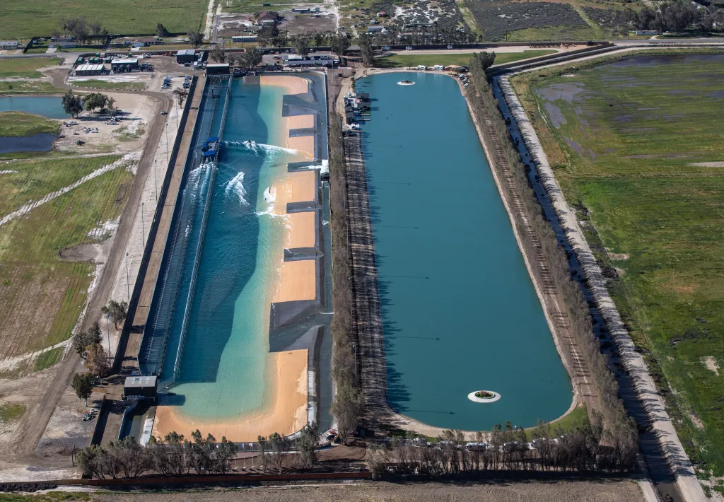 Aerial view of Kelly Slater's Surf Ranch in Lemoore, California, showing two long, artificial lagoons, one with generated waves and a sandy beach-like area, and another calm lagoon, surrounded by agricultural fields and some buildings.