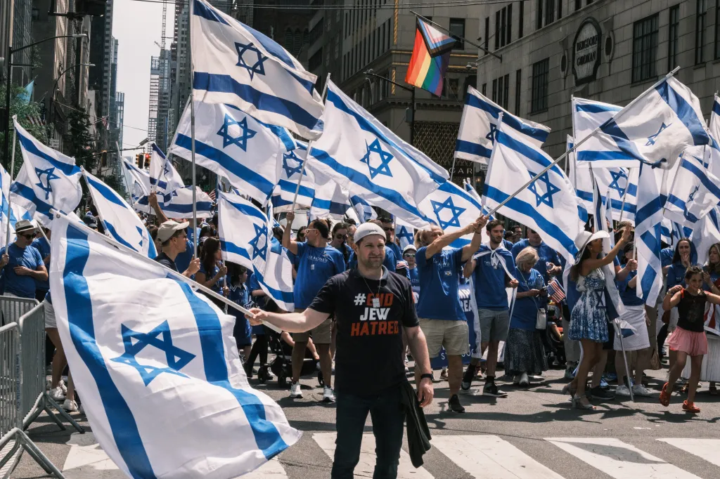 People marching in the Israel Day parade, holding many Israeli flags, with one person in the foreground wearing a 