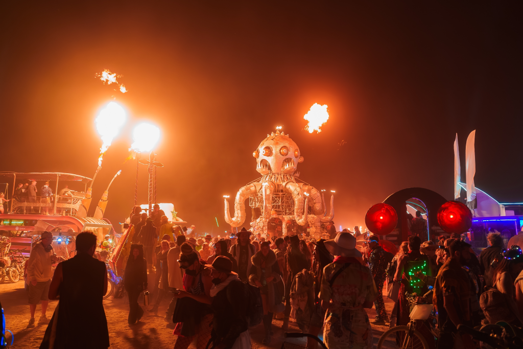 A crowd gathers around a large octopus-shaped art installation with fire effects at the Burning Man festival.