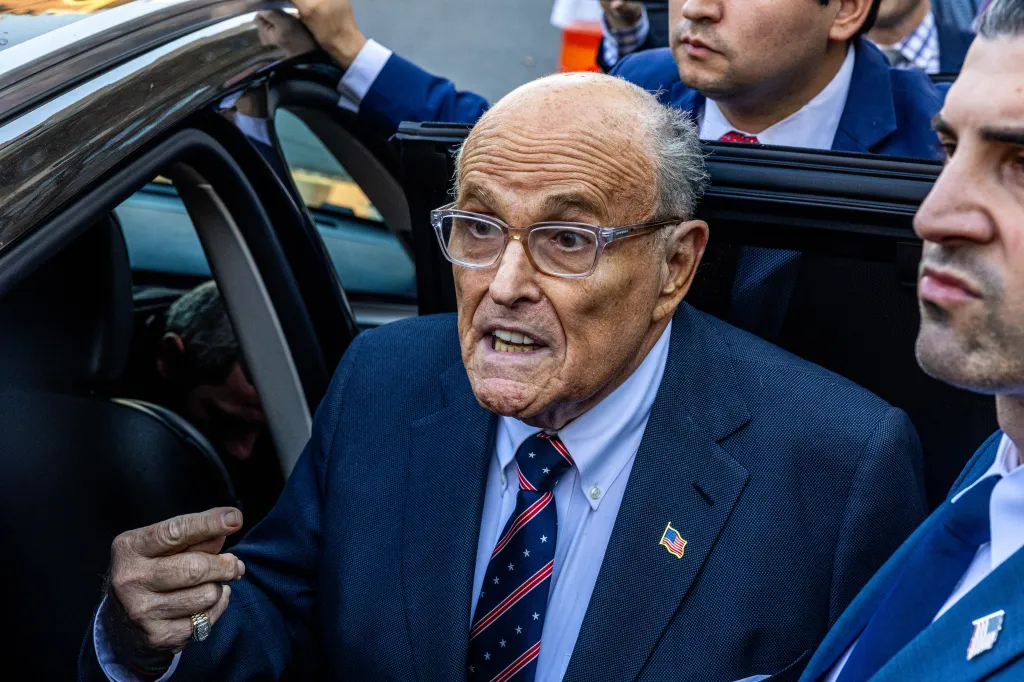 Rudy Giuliani wearing glasses, a suit, and a patriotic tie, exiting a vehicle with a grimace.