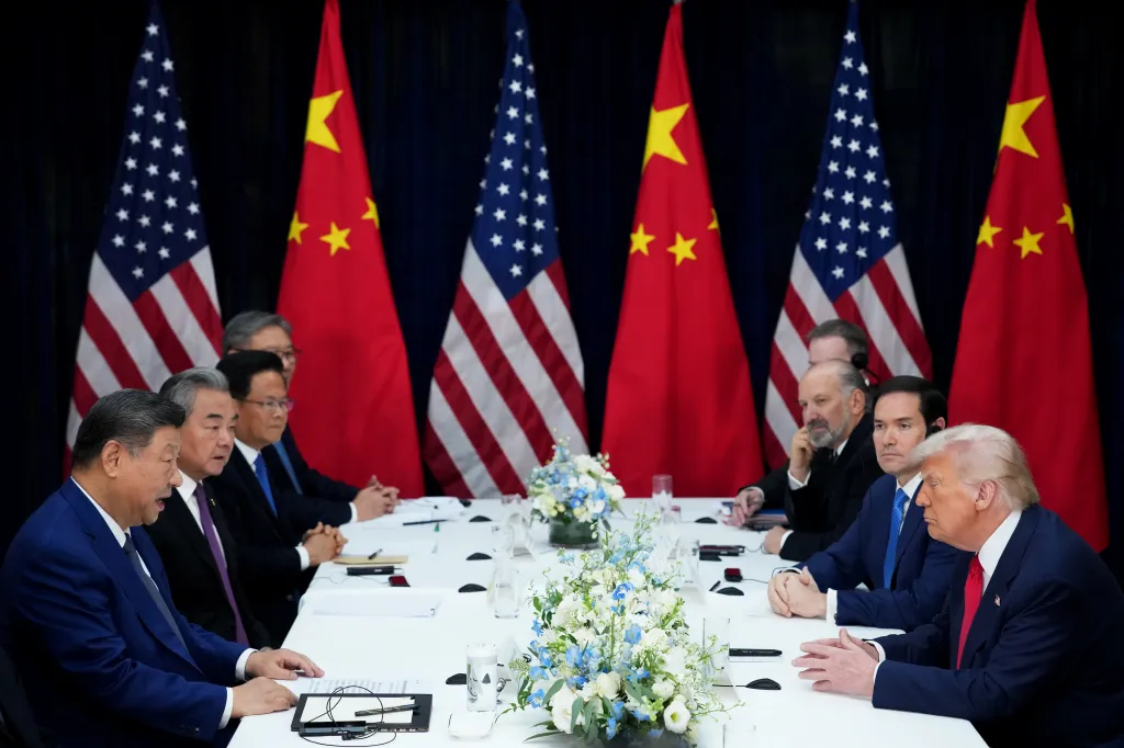 Donald Trump and Xi Jinping meet at a long white table, flanked by US and Chinese flags.