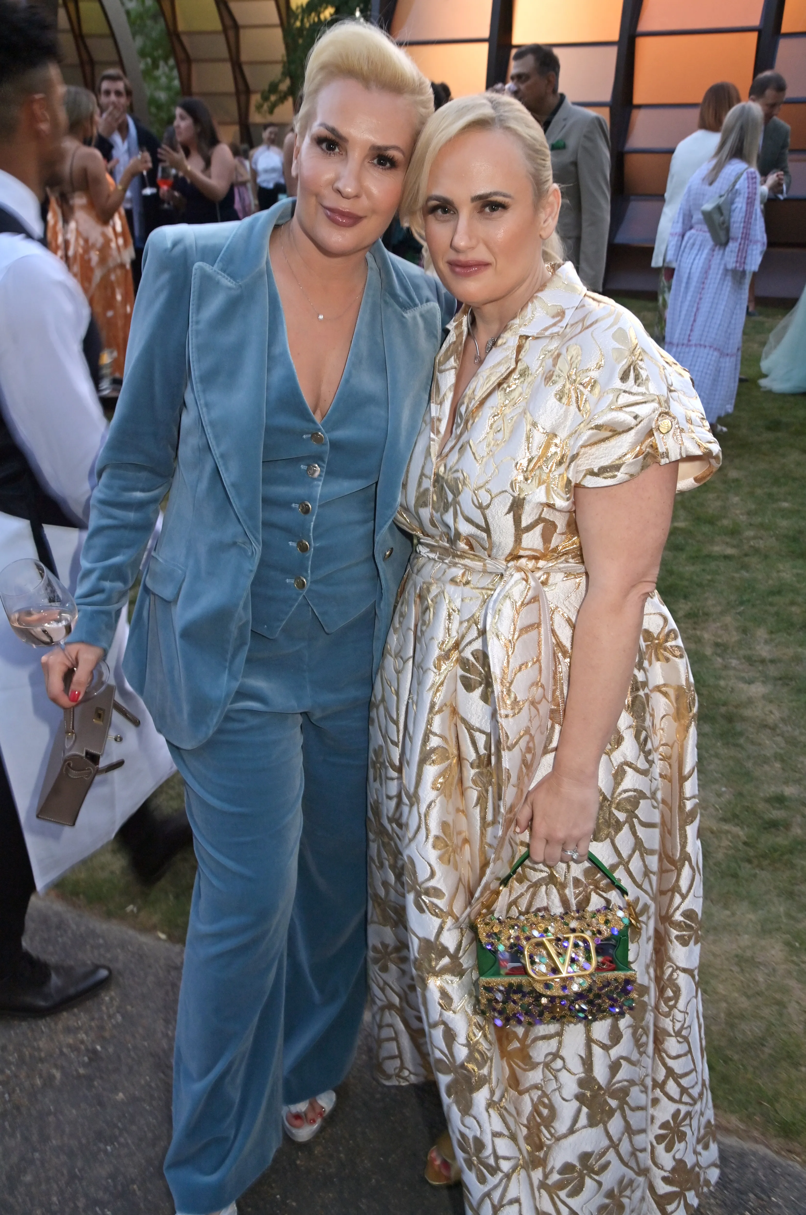 Ramona Agruma in a blue pantsuit and Rebel Wilson in a gold and white floral dress posing together.