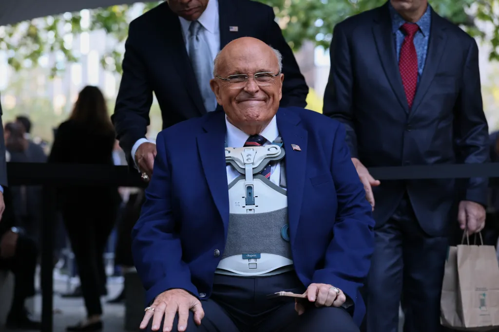 Rudy Giuliani, wearing a blue suit and a medical back brace, smiles while being supported by two men in suits.