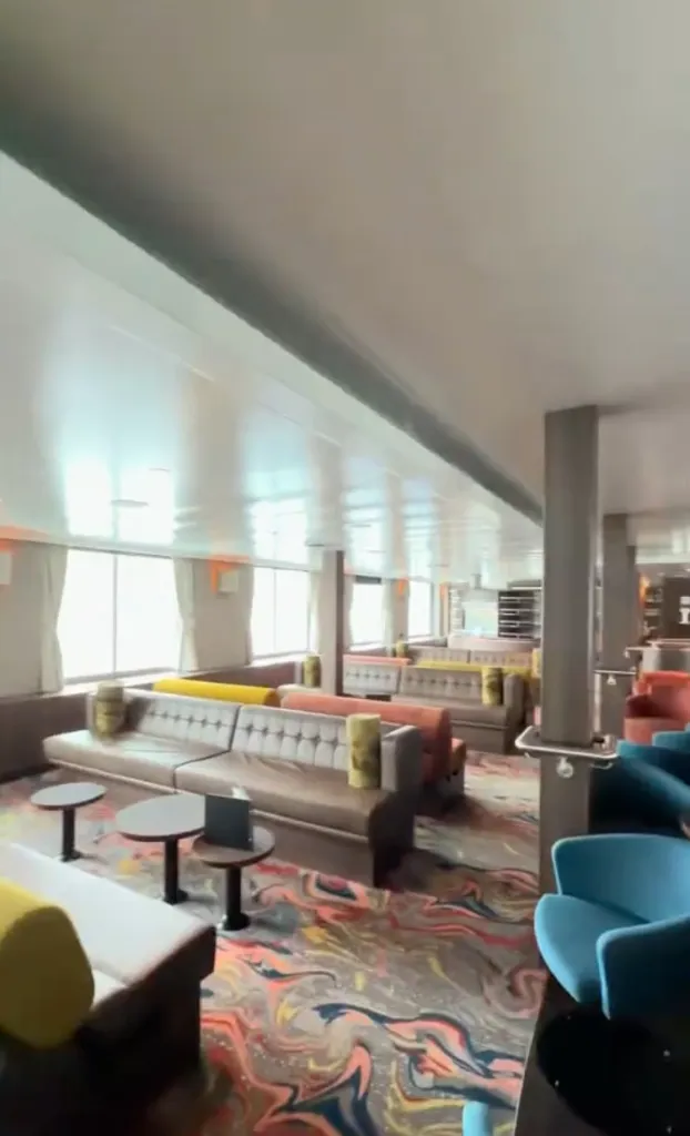 An interior view of the MV Hondius cruise ship lounge area with brown couches, colorful chairs, and a marbled carpet.