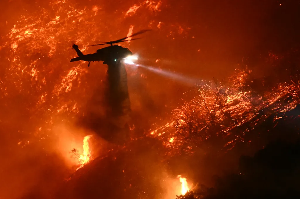 A firefighting helicopter drops water on a wildfire glowing red in the dark.
