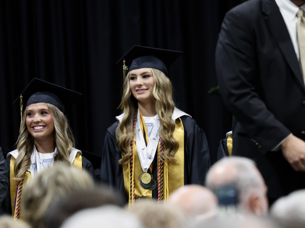 Maddie Aldridge smiling in a black graduation gown and cap, with honors cords and medals, alongside another graduate at a high school graduation ceremony.