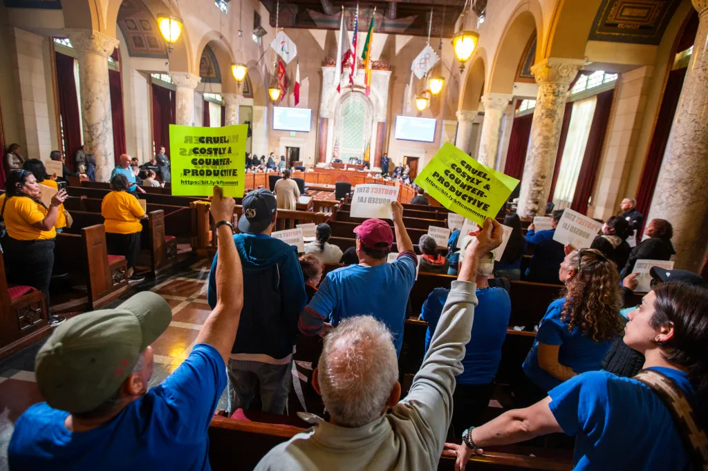 Attendees at a Los Angeles City Council meeting hold signs, with one sign reading 