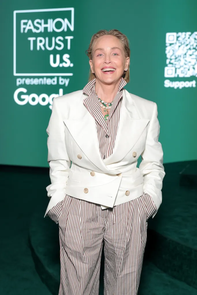 Sharon Stone in a white blazer and brown and white pinstripe shirt and pants.