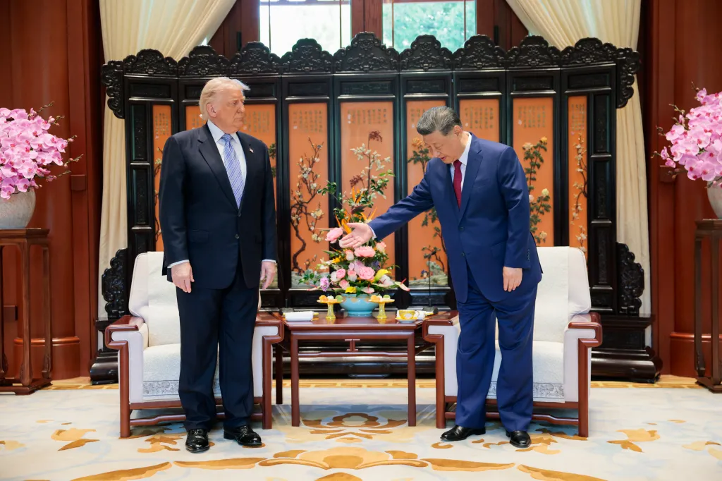 President Trump stands to the left of President Xi, who gestures with his right hand toward a floral arrangement on a table between them.