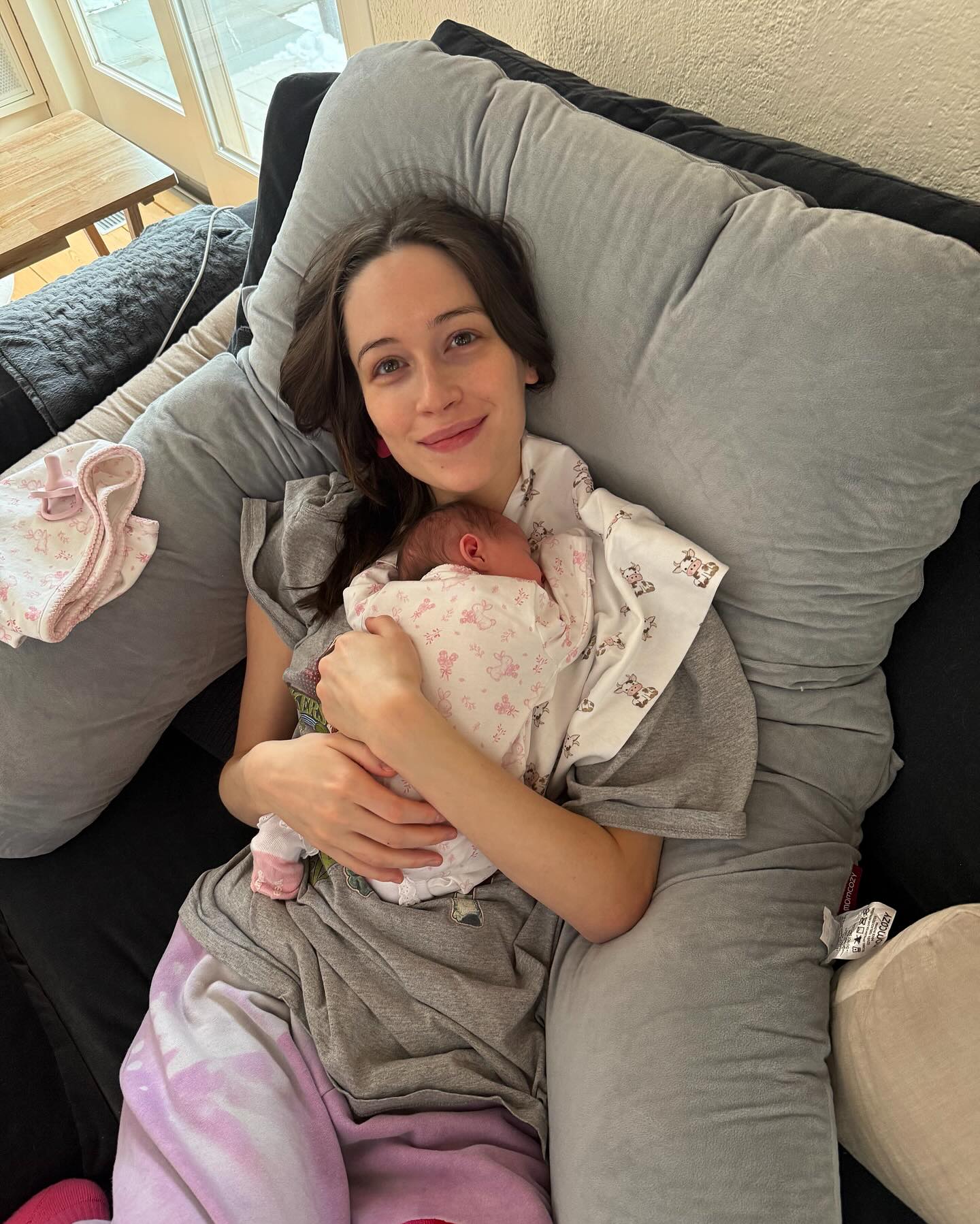 Elsie Hewitt holds her newborn baby close to her chest while lying on a couch with a gray pillow behind her.