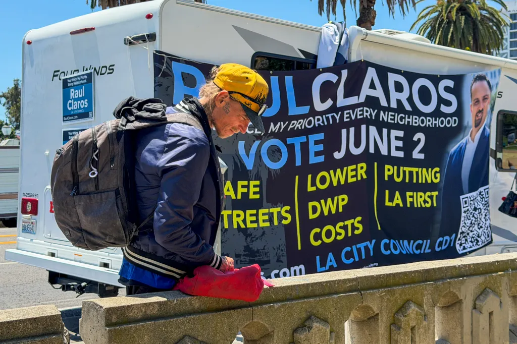 A person wearing a backpack and hat stands before an RV adorned with a large campaign banner for Raúl Claros, Los Angeles City Council candidate.