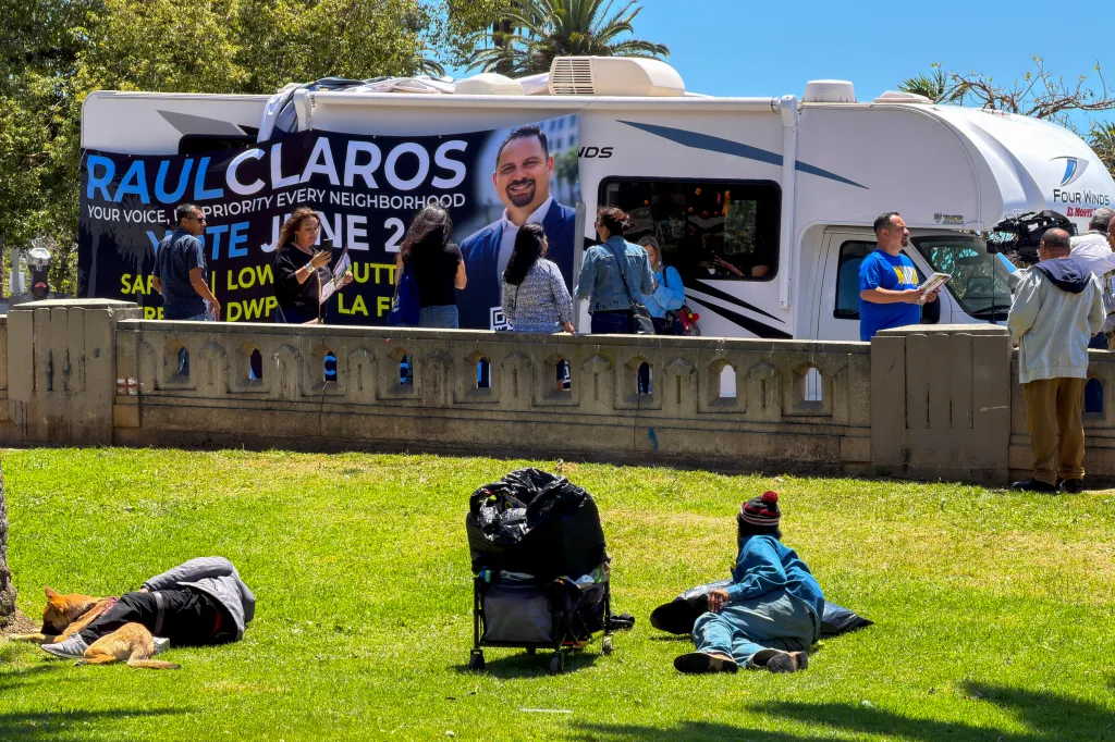 Raúl Claros' campaign RV with a banner and supporters, with several people relaxing on the grass in the foreground.