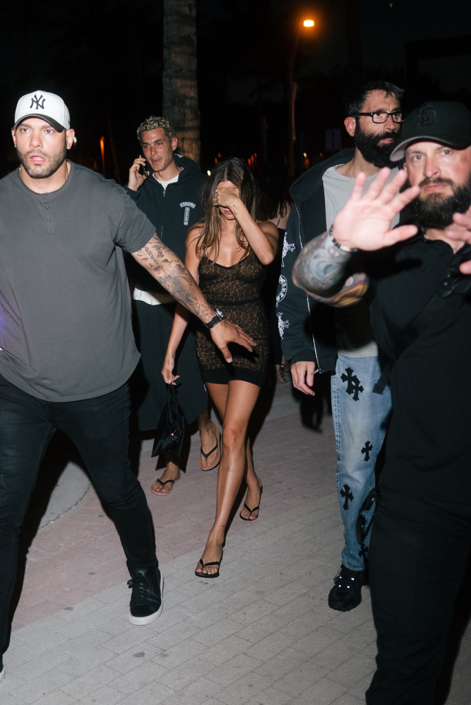 Hailey Bieber in a sheer mini dress covering her face, flanked by security.