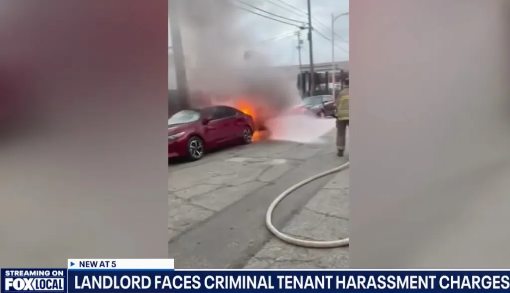 A red car engulfed in flames with white smoke billowing from it, as a firefighter aims a hose at it.