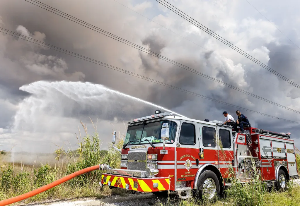 Firefighters spraying water from a fire truck with smoke in the background.