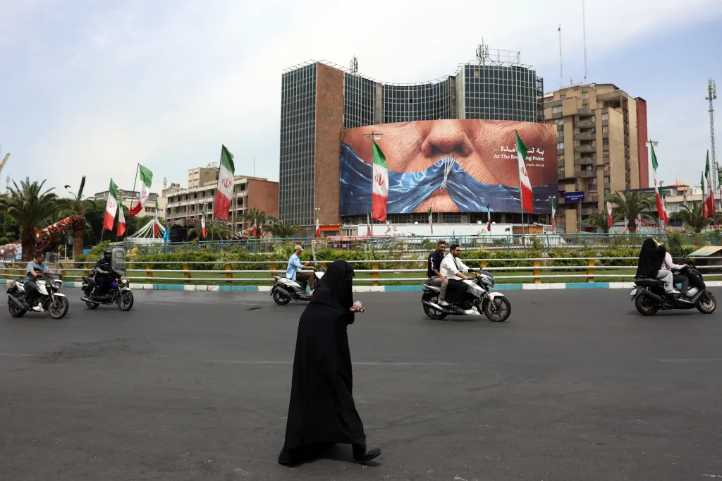 An Iranian woman crosses a road in front of a political billboard of Donald Trump, with Iranian flags on the street.