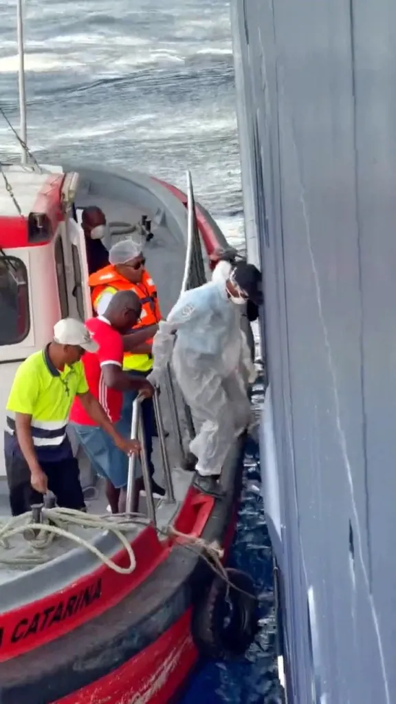 Medical personnel in a hazmat suit boards a cruise ship from a smaller boat.