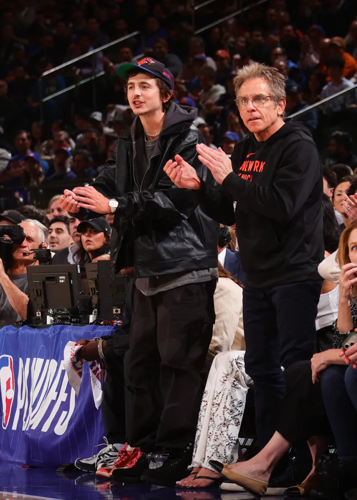 Timothée Chalamet and Ben Stiller watch an NBA game, both wearing casual attire, with Chalamet in a black leather jacket and baseball cap, and Stiller in a black hoodie.