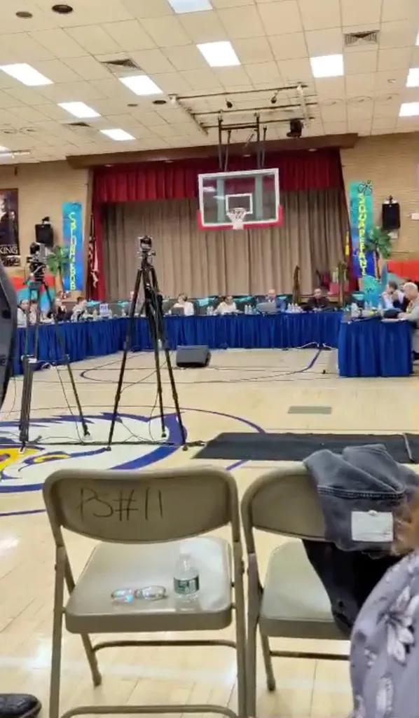 A basketball court set up for a meeting, with a long blue-draped table and a line of people seated behind it.