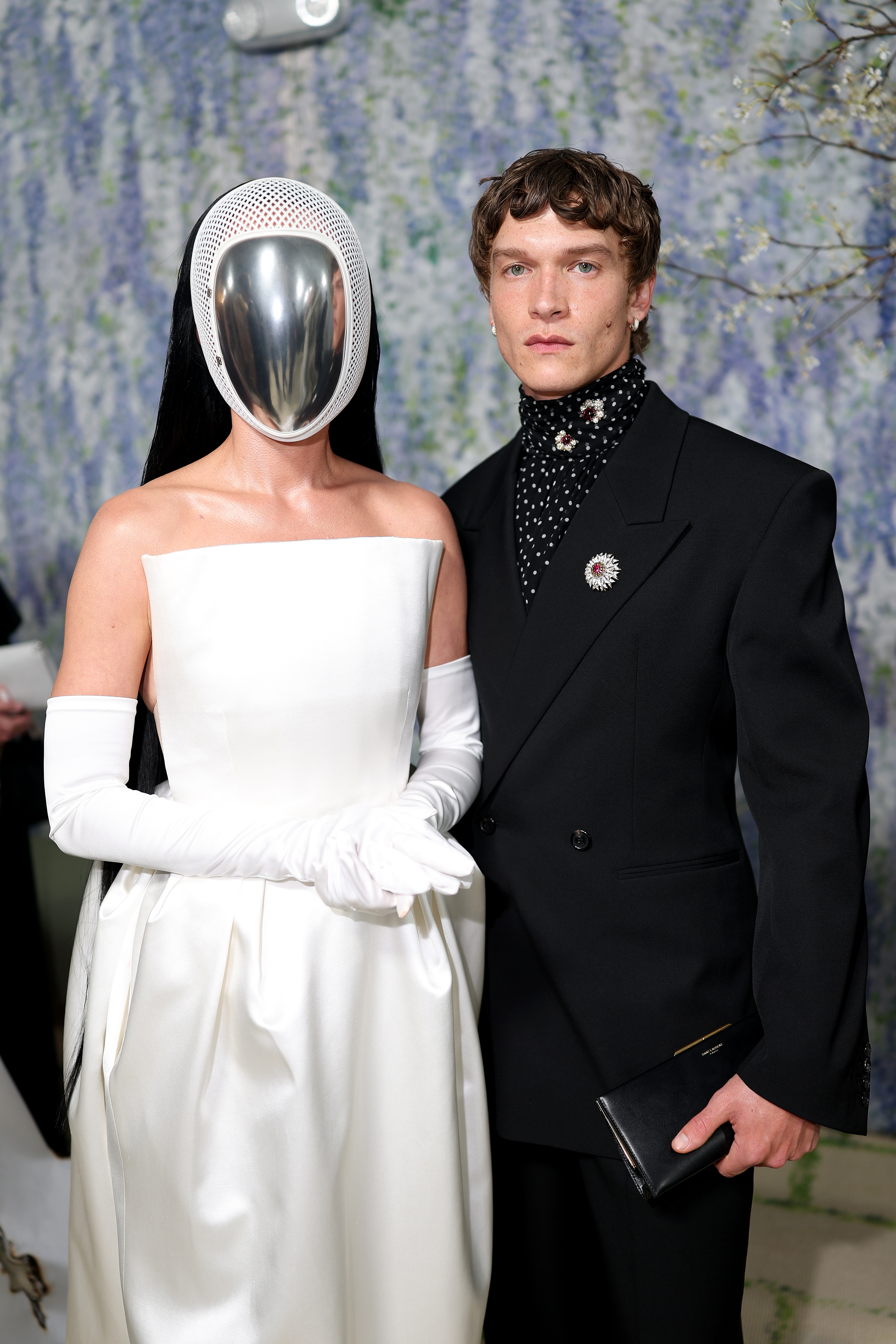 Katy Perry in a white strapless gown and a metallic face mask next to Connor Storrie in a black suit with a polka dot turtleneck, holding a clutch.
