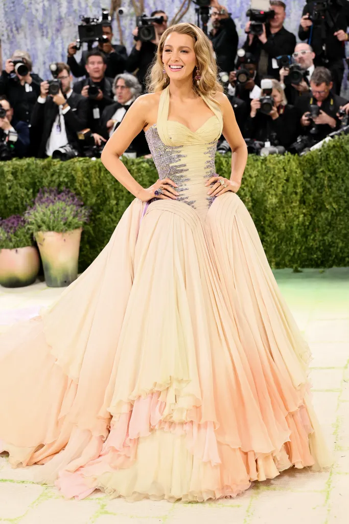 Blake Lively poses in a colorful, flowing gown with photographers in the background.
