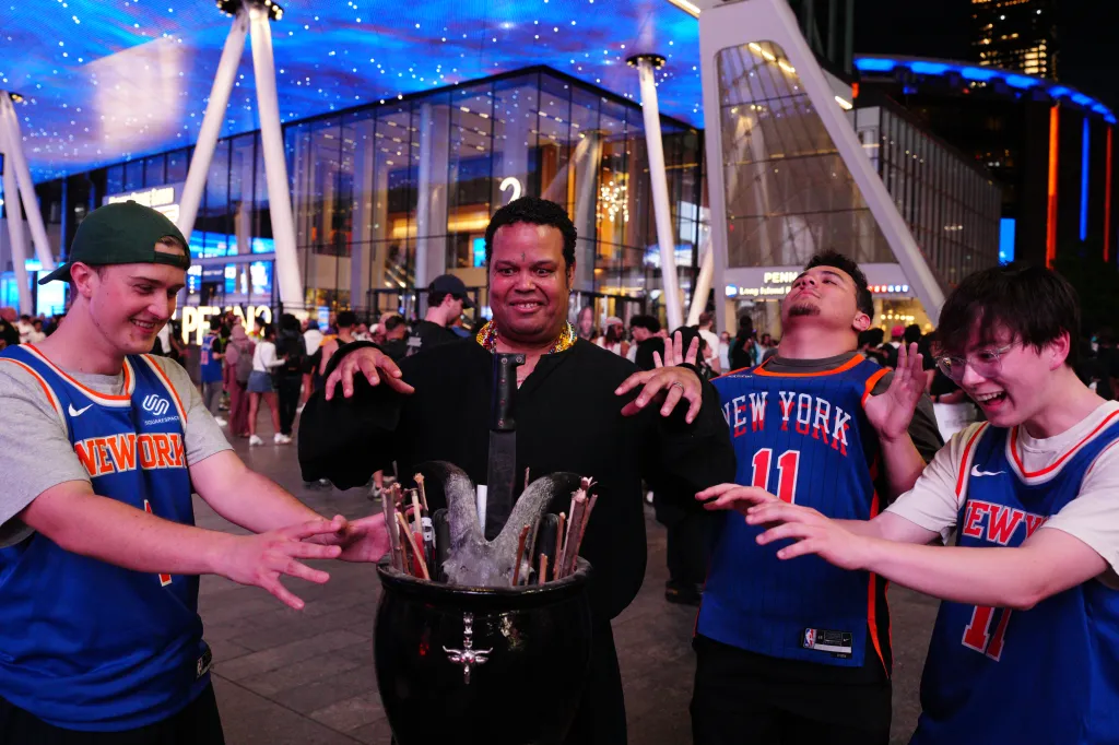 Four men stand around a cauldron with goat horns and a machete, as one man in a black cloak appears to cast a