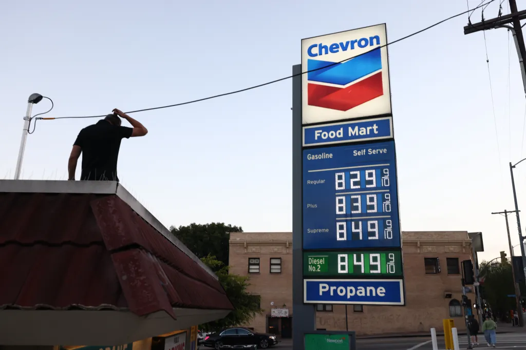 A man scratching his head while looking at a Chevron gas station sign displaying high prices: Regular 8.29, Plus 8.39, Supreme 8.49, Diesel 8.49.