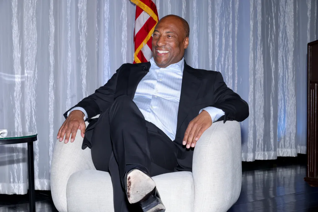 Byron Allen smiling while seated in a chair, with an American flag and white curtains behind him.