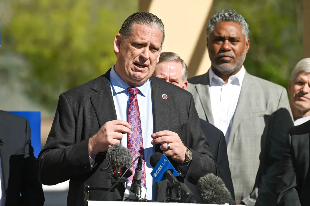 Sen. Tony Strickland speaking at a press conference with microphones in front of him and other men behind him.