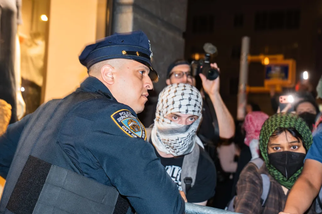 A police officer at an anti-Zionist protest faces a demonstrator with a keffiyeh covering his face, while another protester looks on.