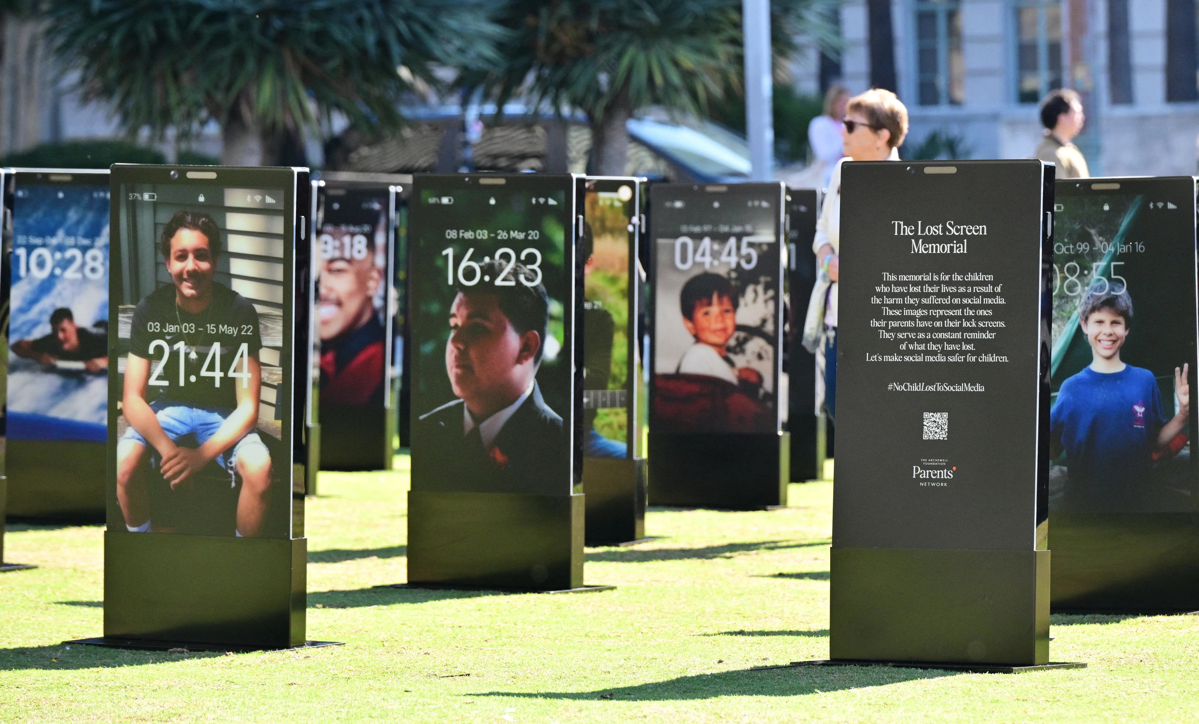 The Lost Screen Memorial features large-scale smartphone displays showing images of deceased children, with one screen displaying text explaining the memorial's purpose in raising awareness about social media harm.