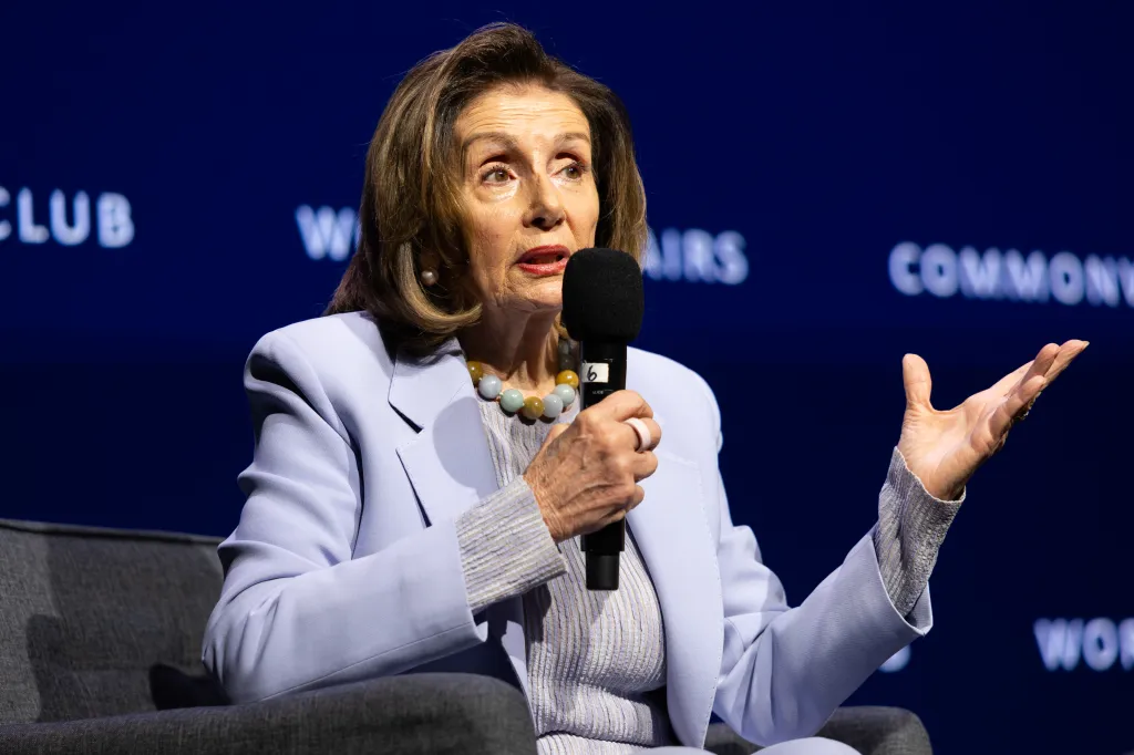 Nancy Pelosi speaking at an event, holding a microphone in her right hand and gesturing with her left hand.