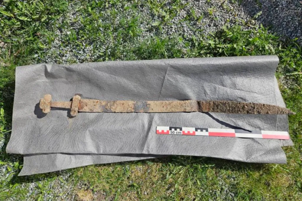 A 1,300-year-old early Viking Age sword, rusty and brown, rests on gray material on grassy ground, with a 50cm measuring stick beside it.