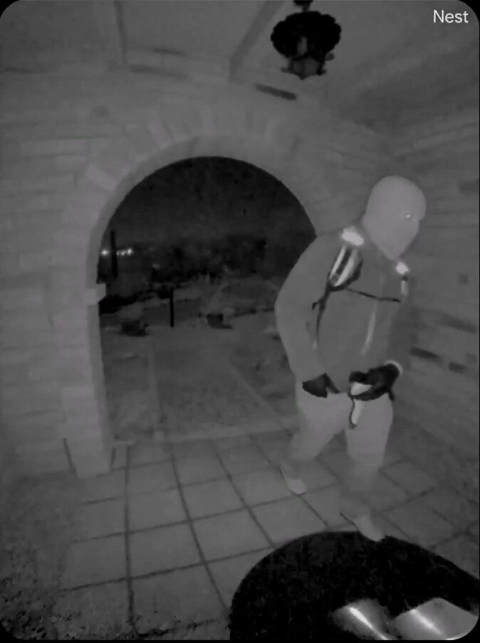 Night vision surveillance image of a person wearing a mask, hoodie, and backpack, walking out of an arched doorway.