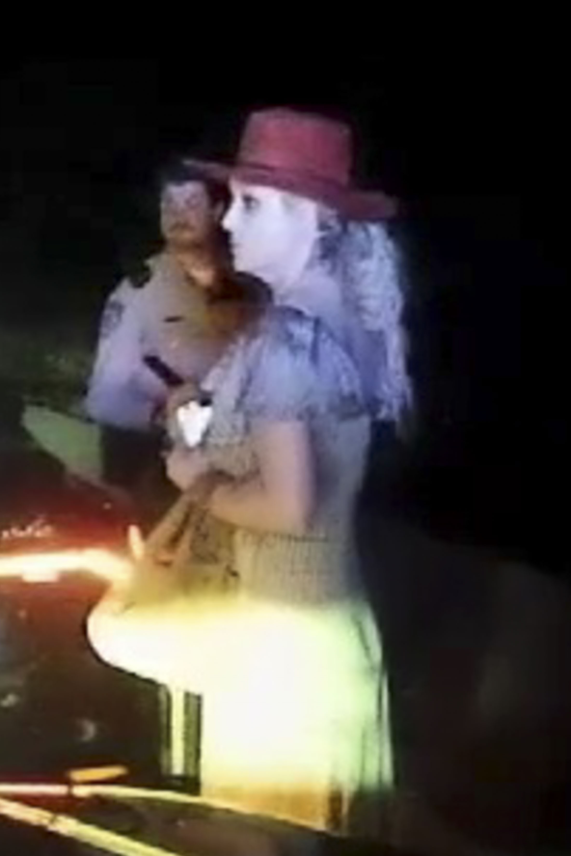 Britney Spears in a red hat being questioned by a police officer at night.