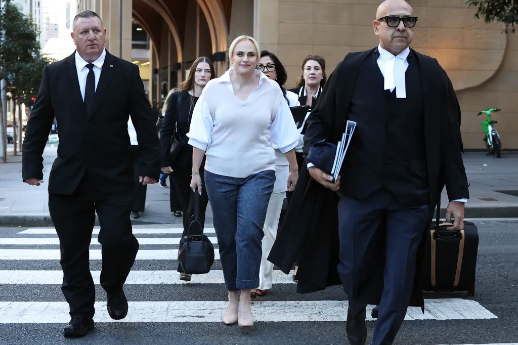 Rebel Wilson walks on a crosswalk surrounded by other people, including two men in suits, heading to court for a defamation trial.