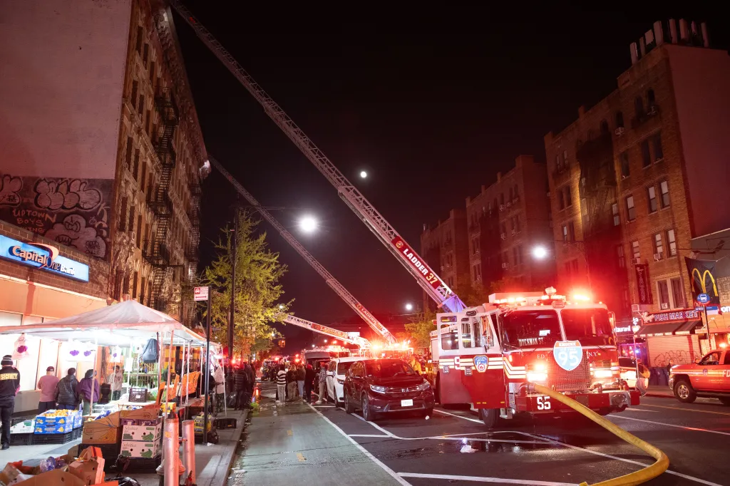 Firetrucks with ladders extended to buildings at night, with people and street vendors on the sidewalk.