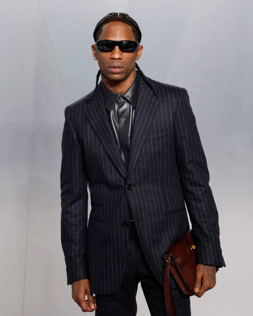 Travis Scott wearing a pinstriped suit, black leather shirt, dark sunglasses, and holding a brown alligator-skin bag.
